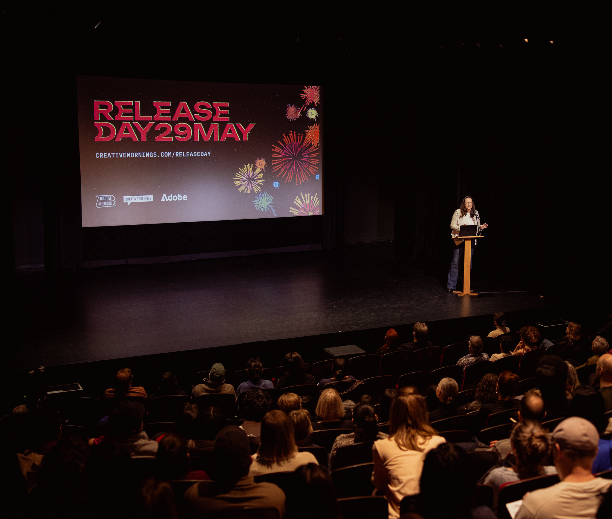 Photo of a speaker stands at a wooden podium on a darkened stage, addressing a seated audience in a theater. A large screen behind her displays bold red text reading 'RELEASE DAY 29 MAY' alongside the URL creativemornings.com/releaseday, decorated with colorful firework graphics in pink, orange, yellow, and blue. Logos for CreativeQuests, CreativeMornings, and Adobe appear at the bottom of the screen. The audience is visible from behind, watching attentively.