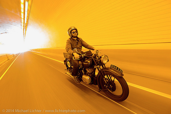 A man wearing a helmet and sunglasses rides a vintage motorcycle through a tunnel, bathed in warm yellow light, with motion blur emphasizing speed and forward movement.
