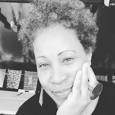 Black-and-white close-up portrait of a woman with short curly hair resting her face on her hand, wearing a large statement ring and a long earring, with artwork pinned on the wall behind her.