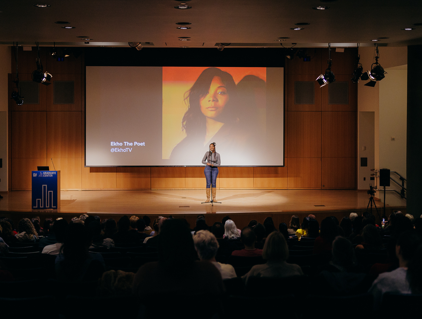 Photo of woman standing on stage speaking into a standing microphone. On a large projection screen behind her is a headshot of herself. Several rows of an audience are in the foreground.