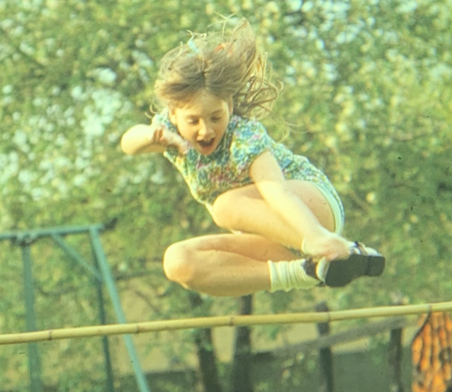Amy, as a little girl wearing a floral jumper and mary jane shoes, is jumping over a bamboo pole. The sun is shining and the grass and trees are lush and green with summer energy.