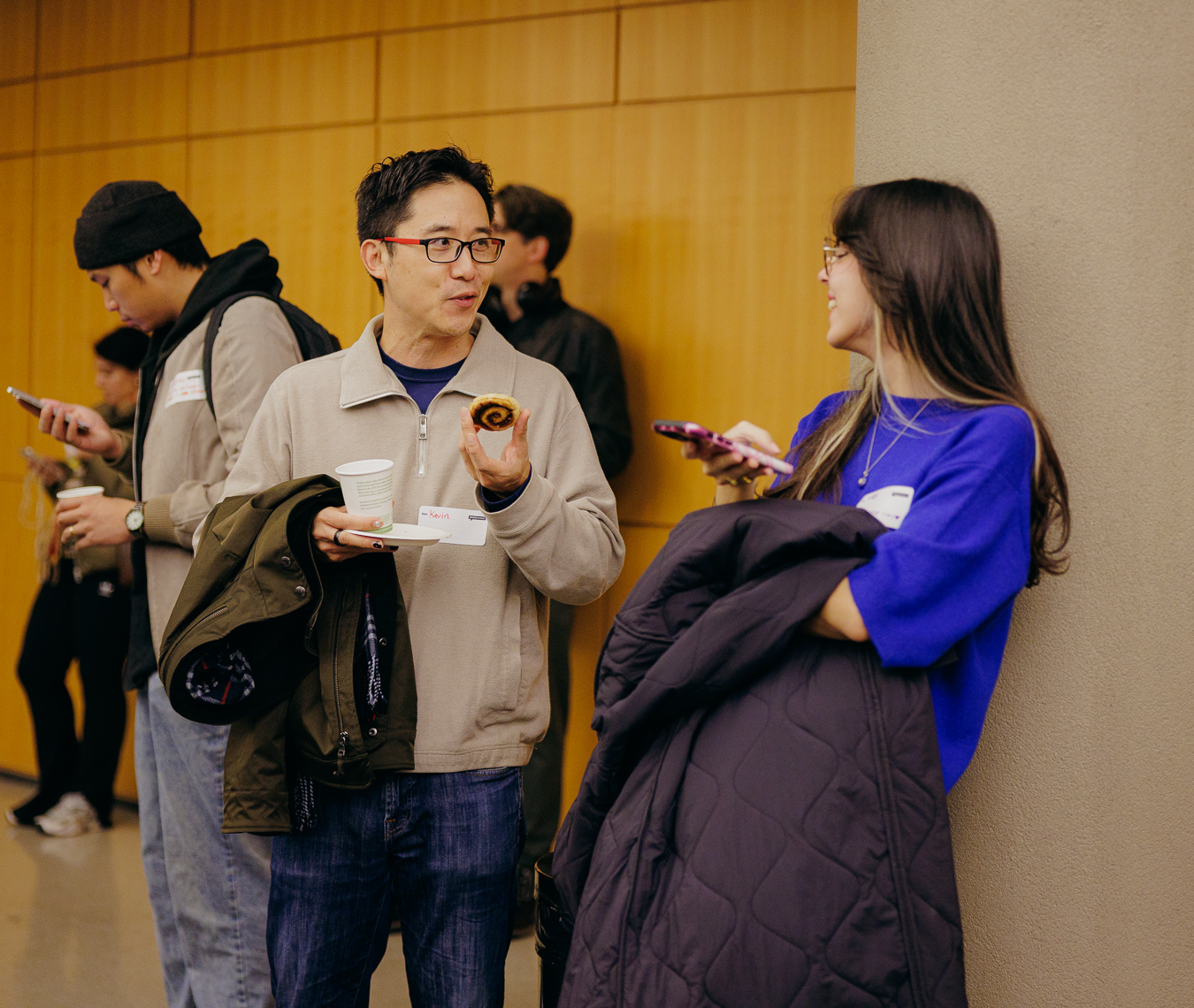 Image of a man holding a paper cup of coffee with glasses, brown jacket and blue jeans talking to a woman wearing a blue sweating and holding a large puffy coat folded across one arm.