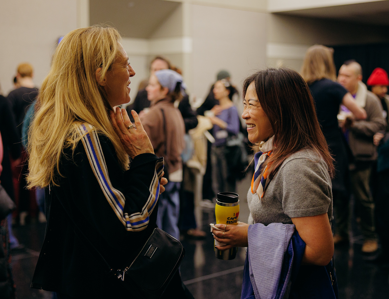 A woman with long, blonde hair wearing a black sweater with blue stripes is talking to a woman holding a coffee mug and wearing a grey sweater with a blue silk scarf.