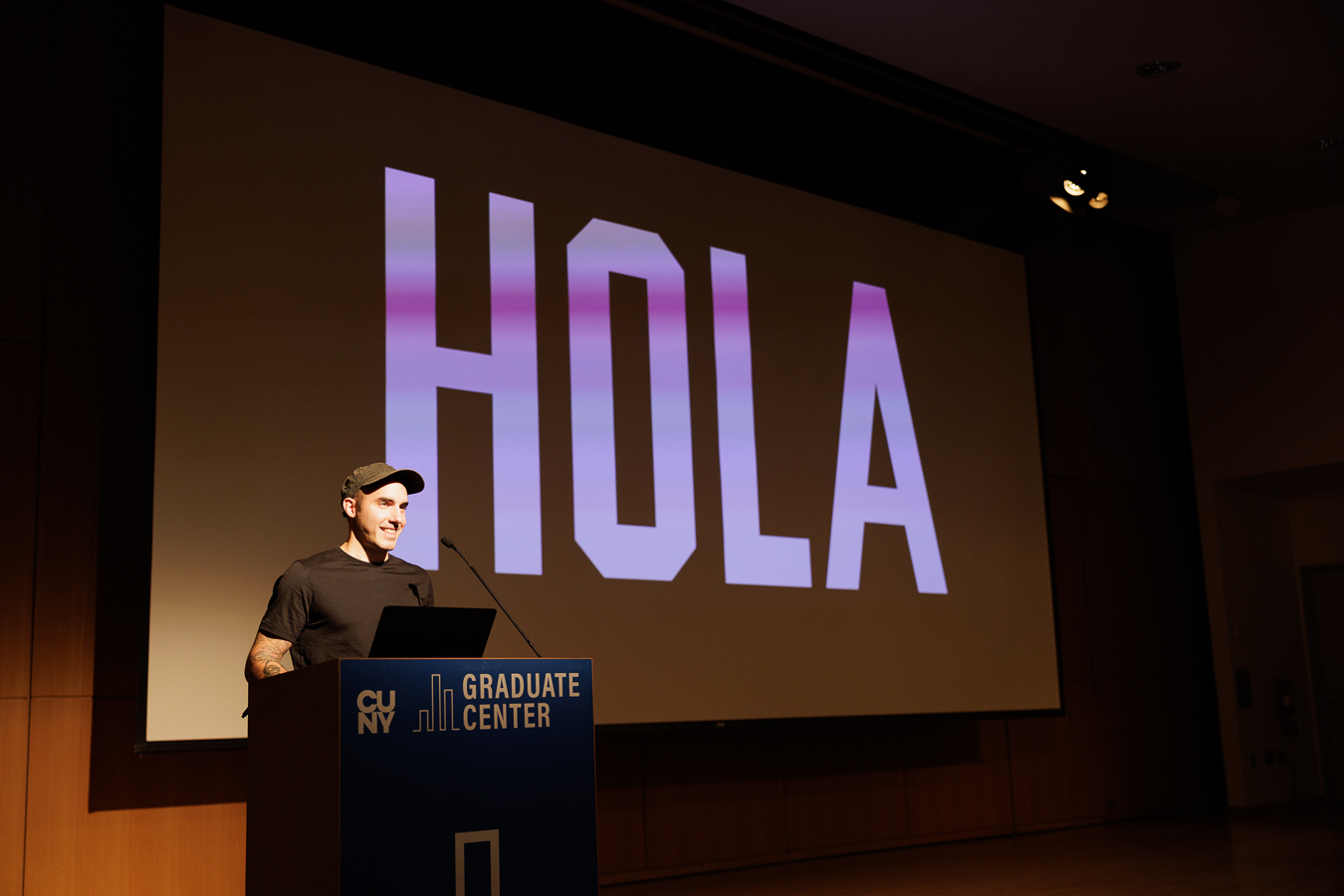 Photo of a man wearing a black tee shirt and black cap standing behind a podium with a small microphone attached. There is a large projection screen behind him with the word hola displayed in block type.