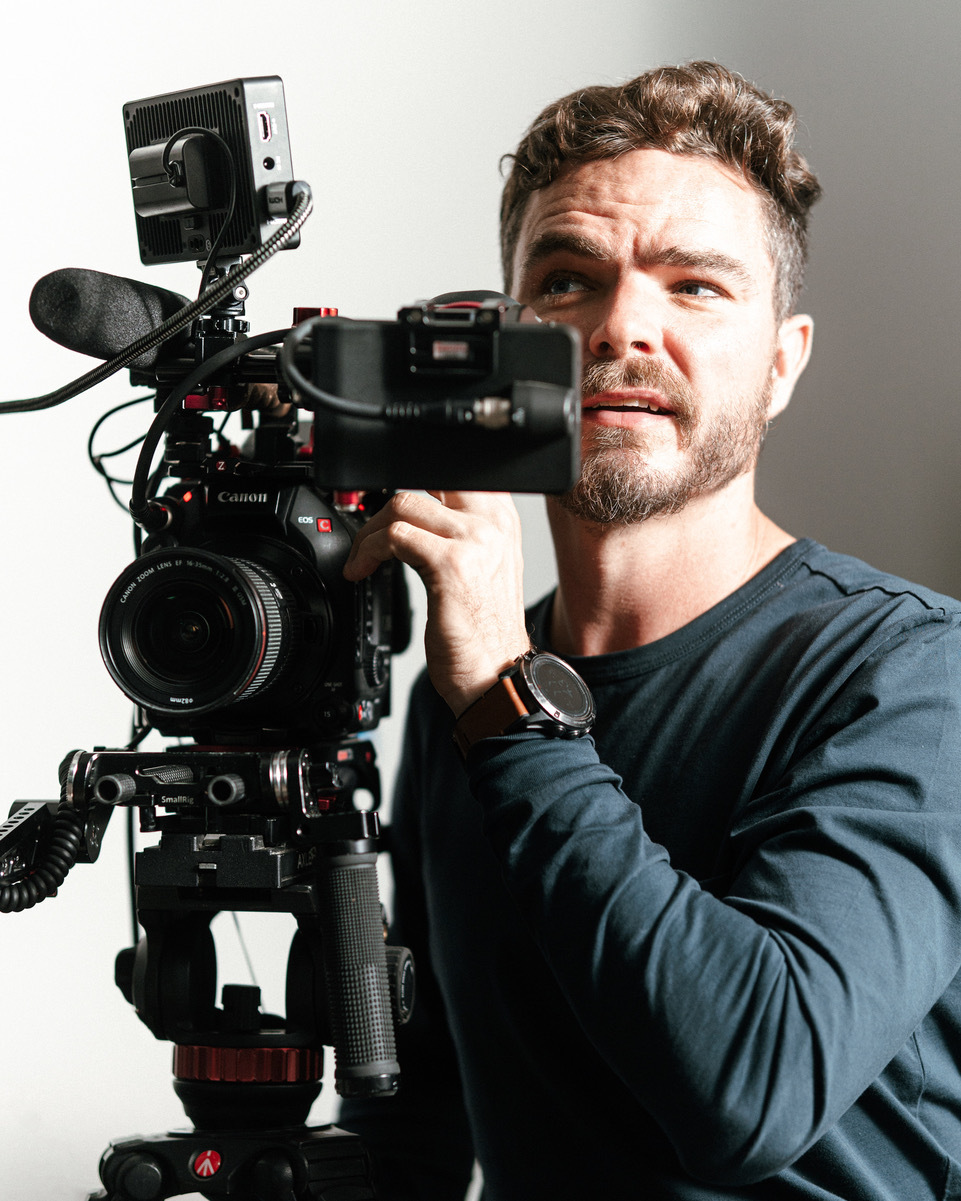 Photo of a man with short curly hair and a beard operates a professional Canon video camera mounted on a tripod. He looks slightly upward and to the side while adjusting the camera, lit by bright natural light.